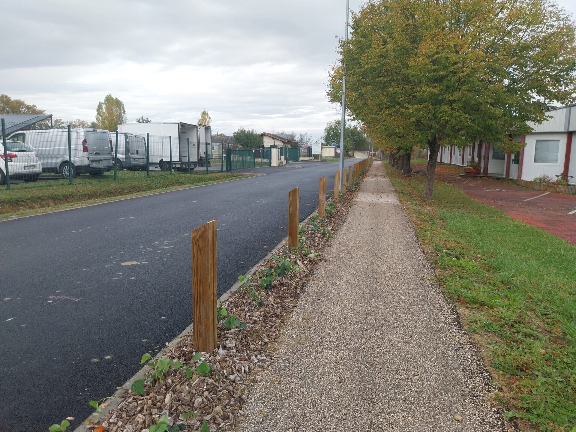 Trottoir avec poteaux bois, paillis et jeunes plantes longeant une route goudronnée.