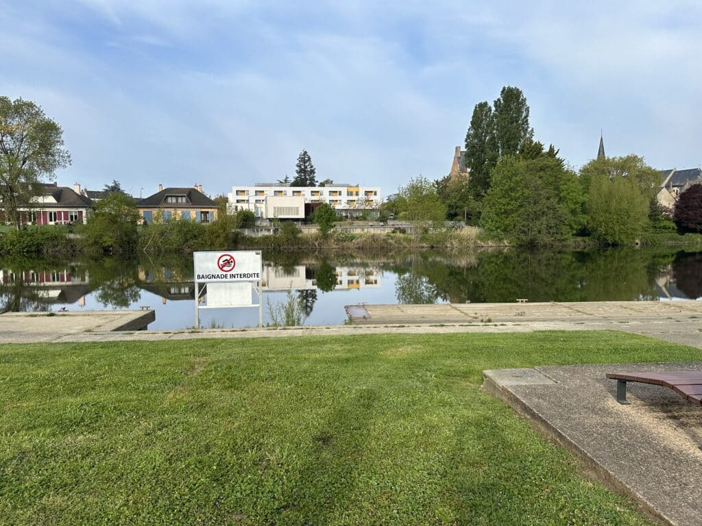 Panneau BAIGNADE INTERDITE devant une rivière calme, maisons et arbres en arrière-plan.