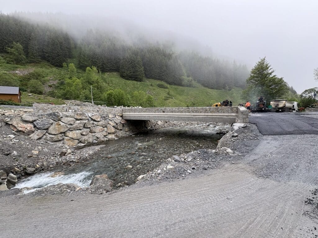 Pont en pierre sur torrent de montagne, travaux d'asphaltage et forêt sous la brume.