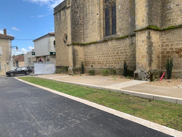 Église ancienne en pierre bordée par une route neuve, une pelouse et un trottoir sous un ciel bleu.