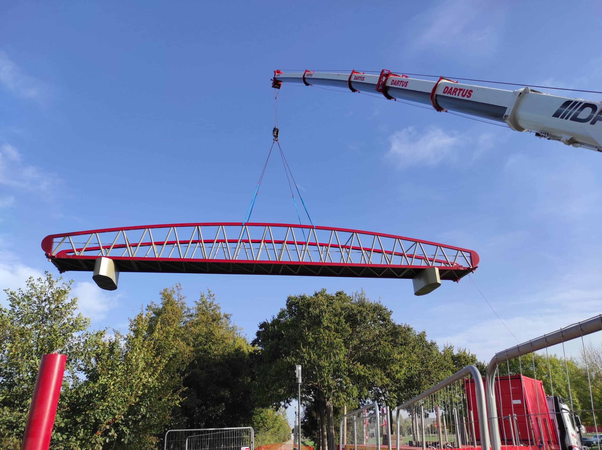 Grue Dartus levant une passerelle rouge métallique au-dessus d'arbres sous un ciel bleu.