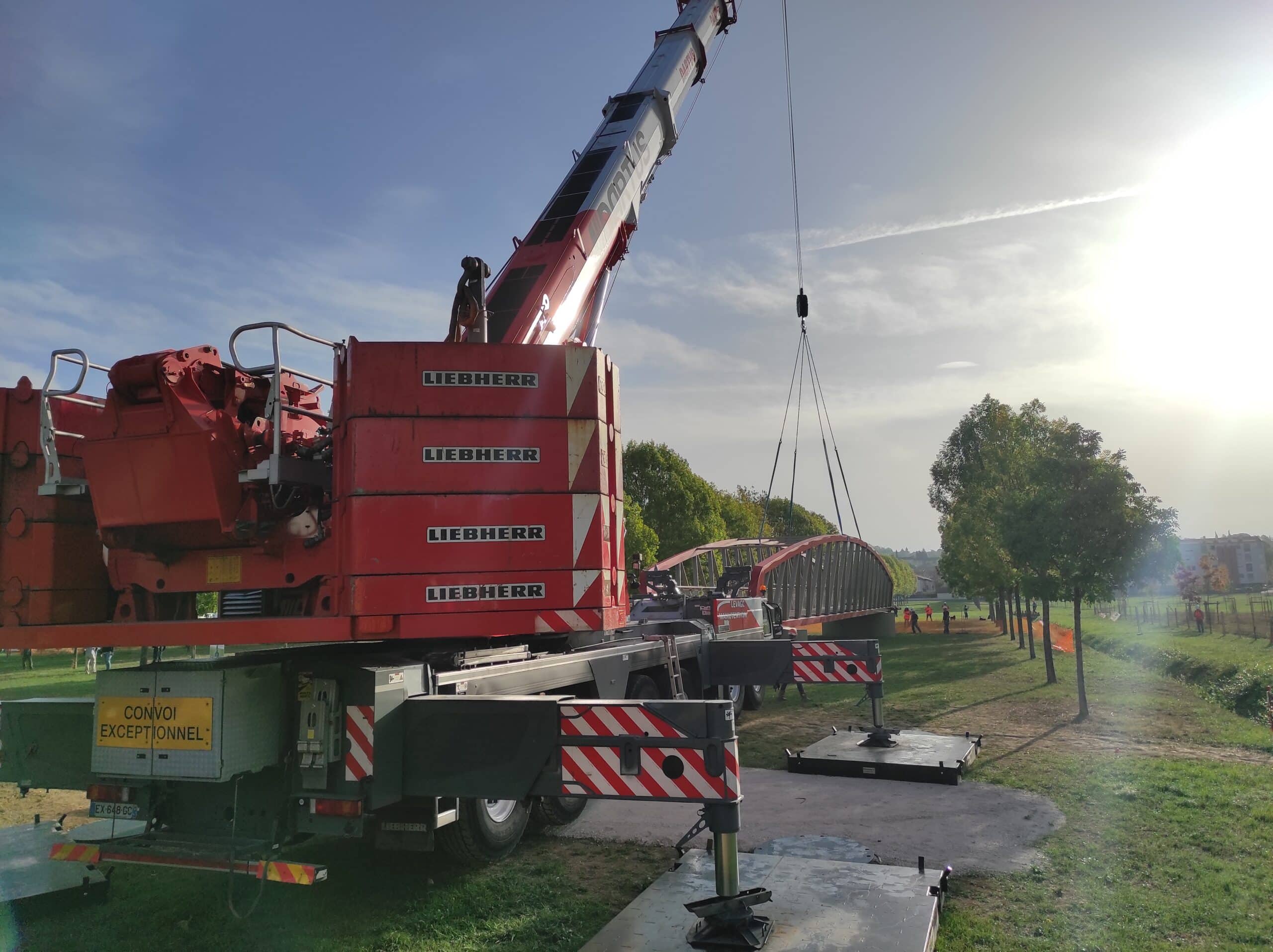 Grue Liebherr rouge installant un pont métallique dans un parc. Marquage CONVOI EXCEPTIONNEL.