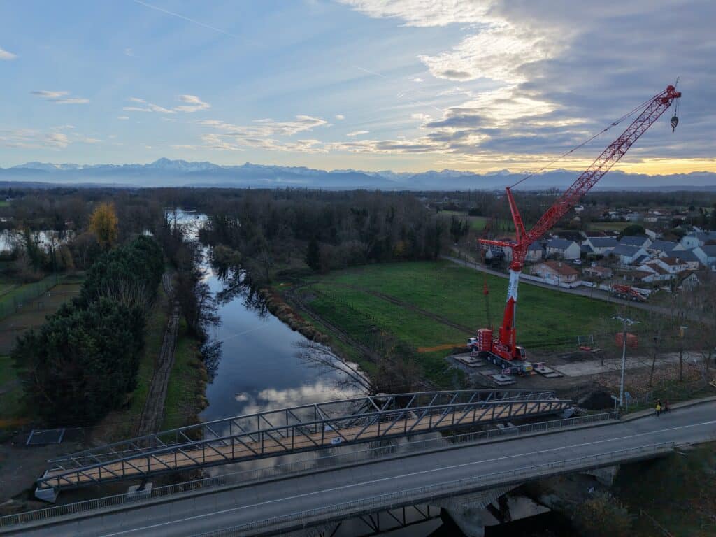 Grue rouge assemblant une passerelle métallique sur une rivière devant la chaîne des Pyrénées au crépuscule.