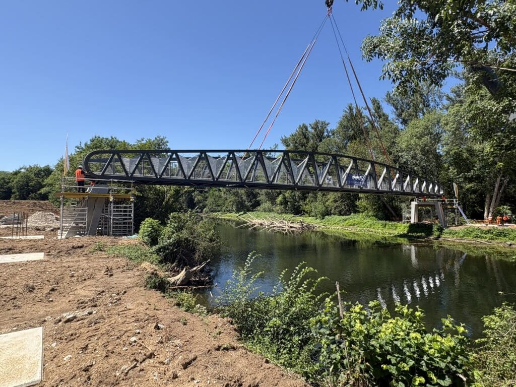 Grue installant une passerelle métallique en treillis au-dessus d'une rivière sur un chantier de construction.
