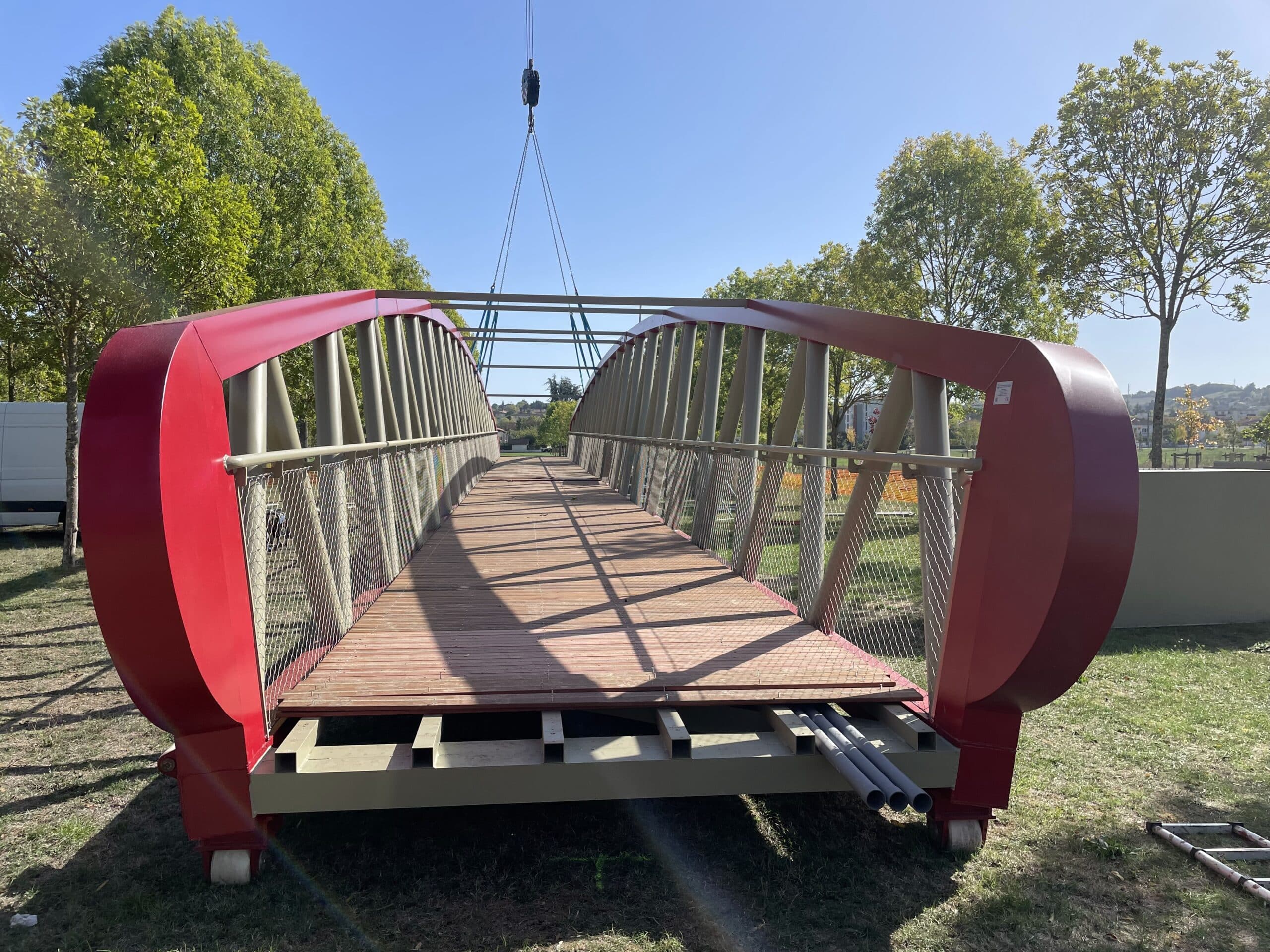 Passerelle piétonne moderne rouge et bois soulevée par une grue dans un parc verdoyant.