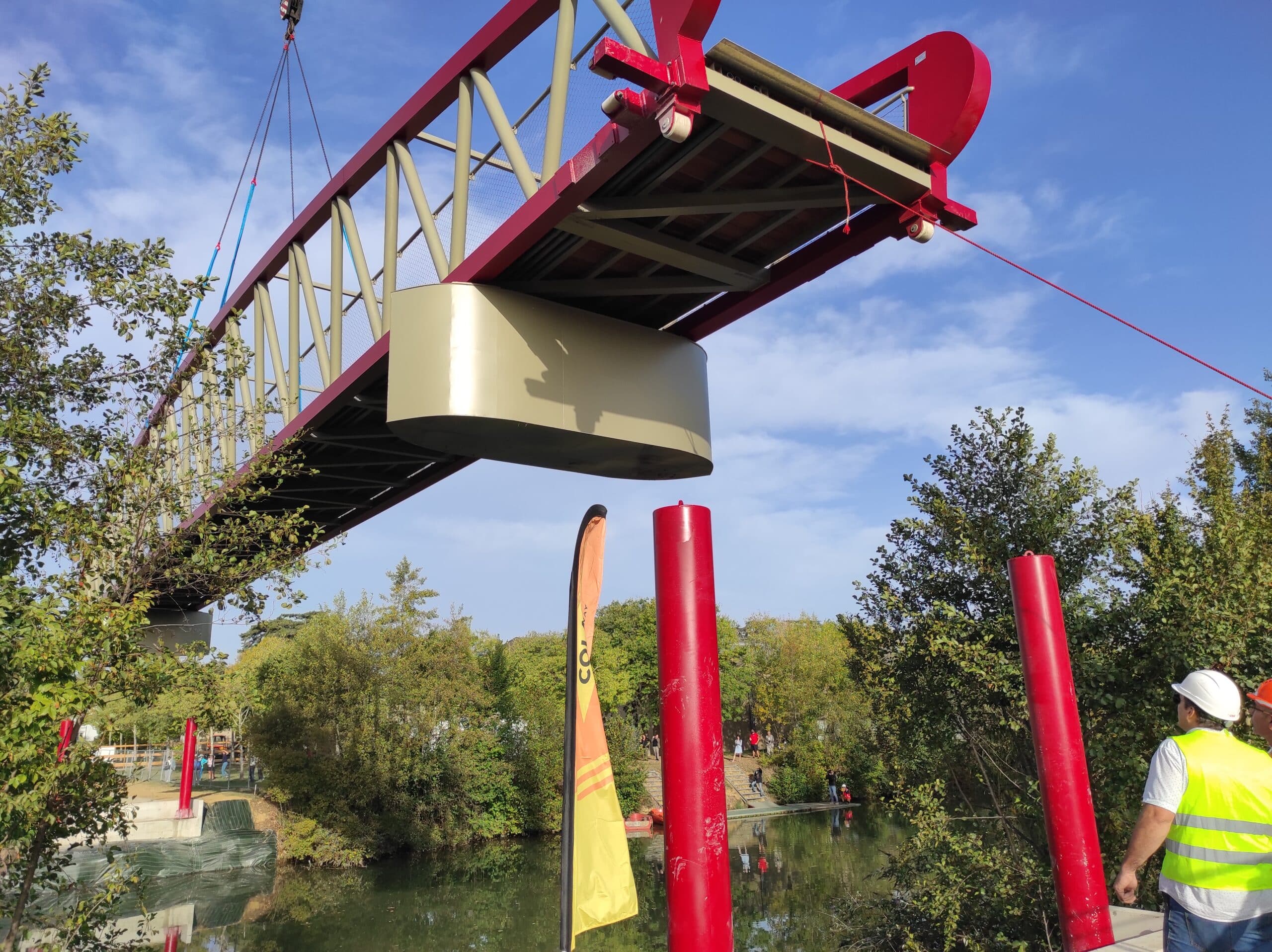 Installation d'une passerelle rouge par grue au-dessus d'une rivière, ouvrier surveillant le chantier.