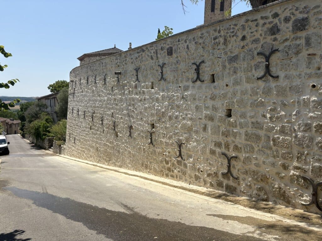 Mur en pierre avec ancres de façade métalliques longeant une rue de village.