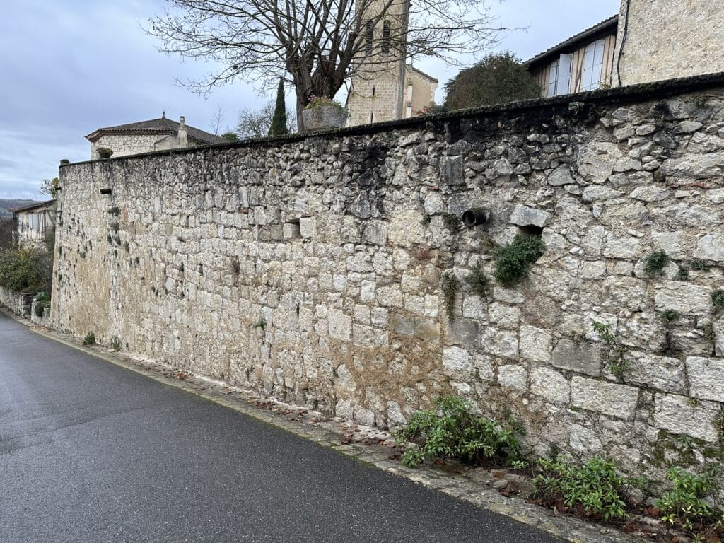 Ancien mur de pierre bordant une route de village français avec clocher d'église et maisons en arrière-plan.