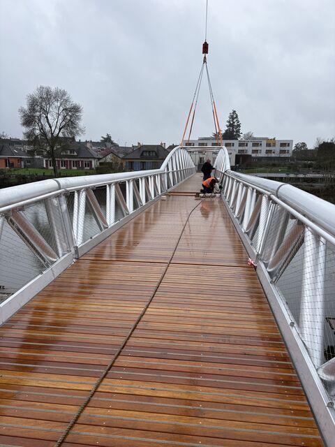Installation de passerelle piétonne bois et métal par grue avec ouvriers en plein chantier.