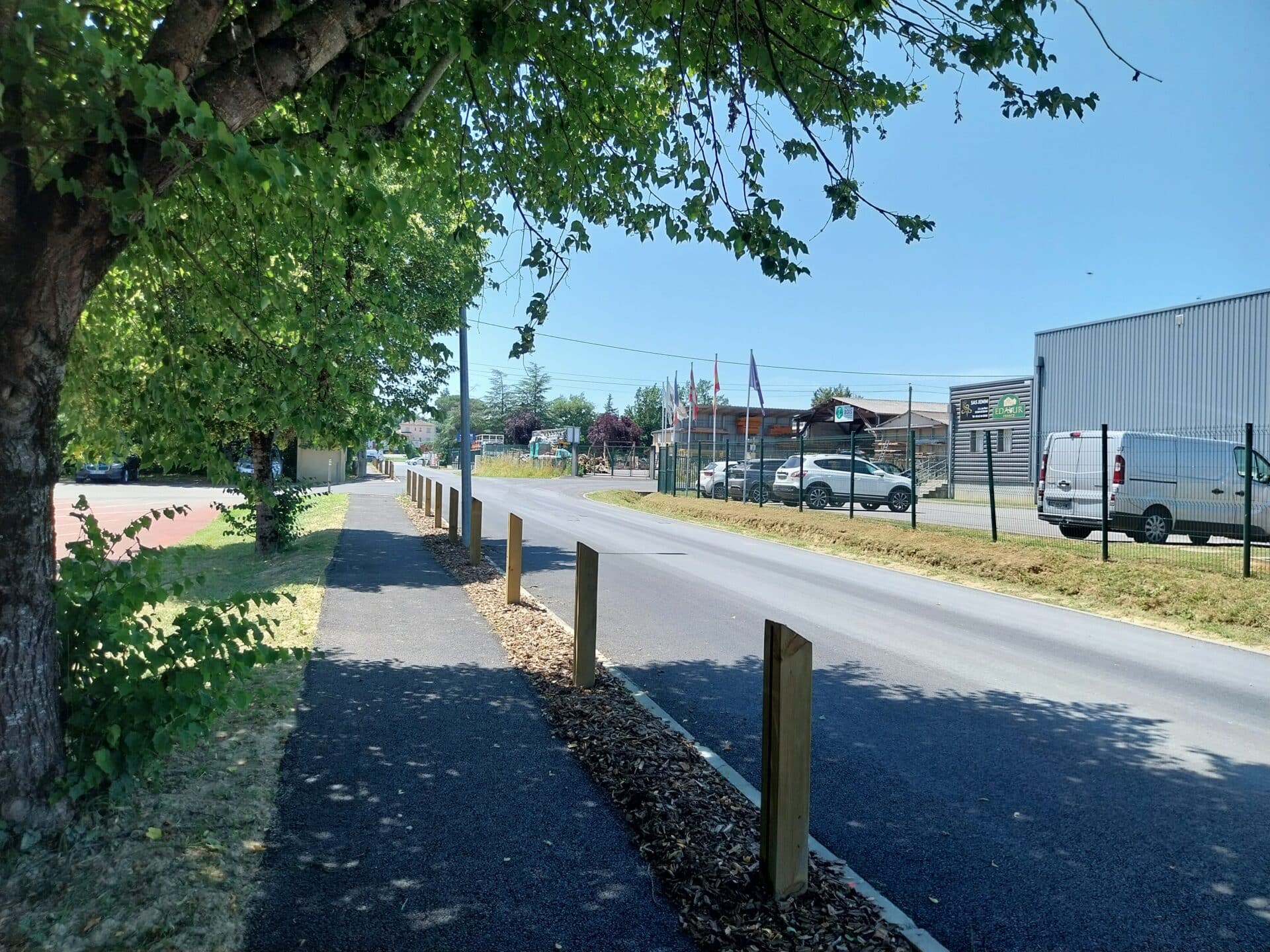 Route avec piste cyclable, poteaux bois, zone industrielle SAS JENN EDAKUR et arbres sous un ciel bleu.