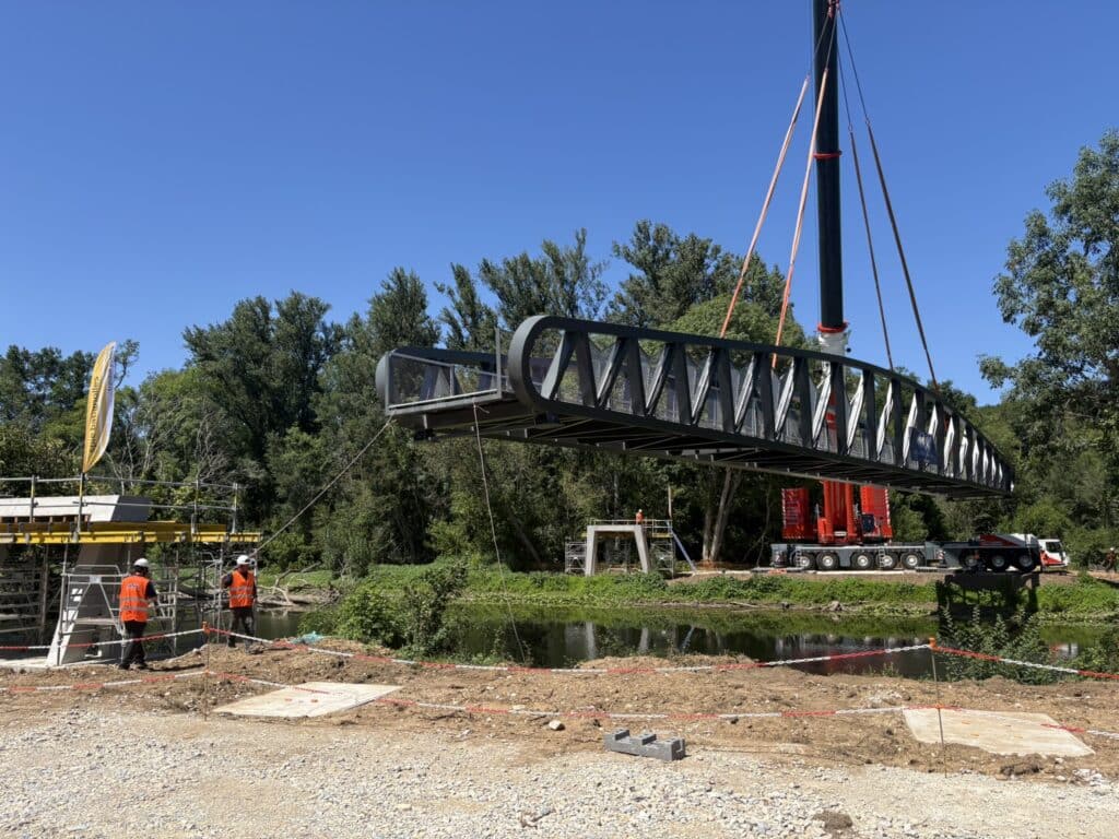 Grue levant une passerelle métallique noire Spie Batignolles sur un chantier fluvial avec ouvriers.