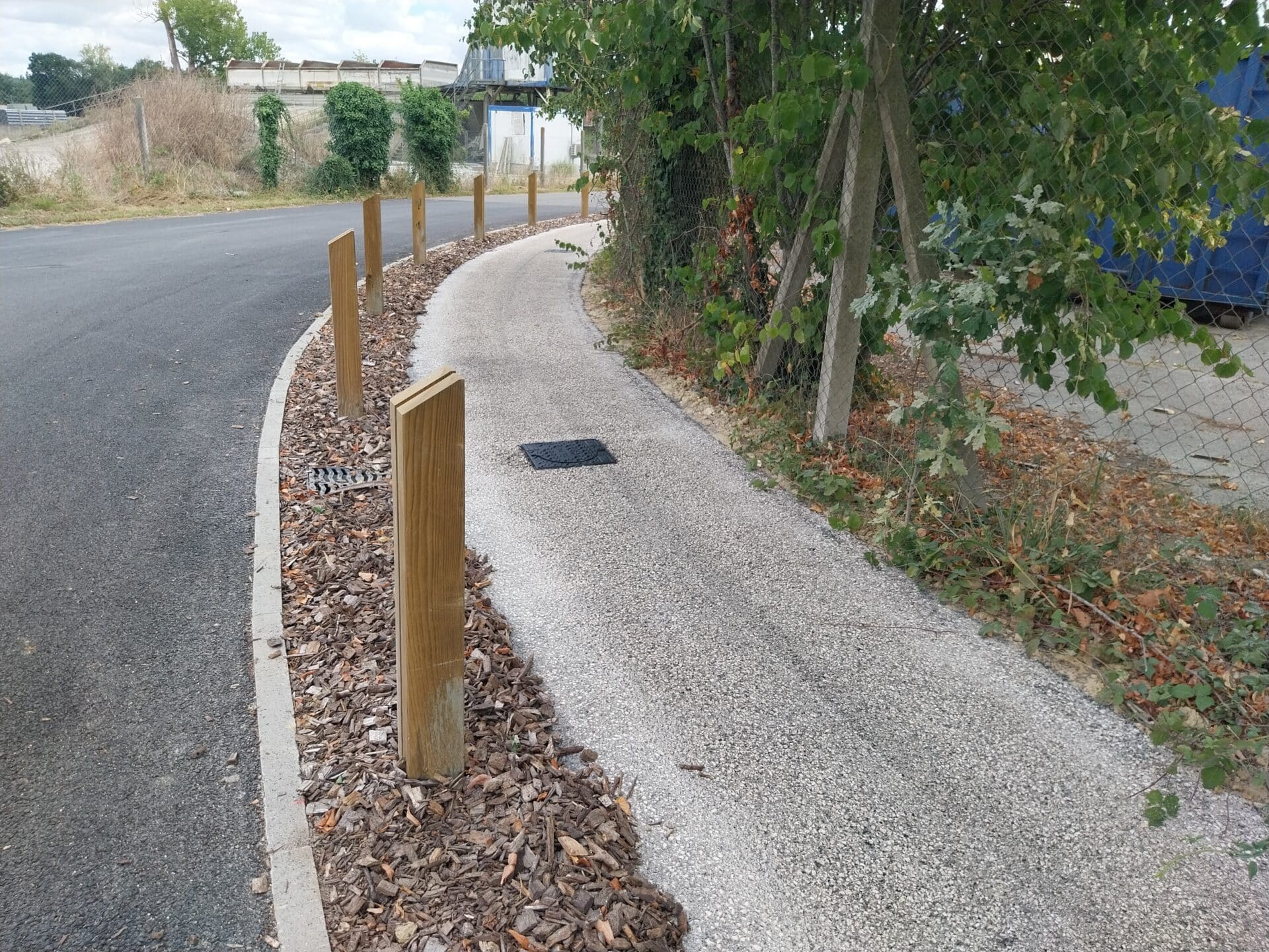Sentier de gravier avec poteaux bois et paillis d'écorce longeant une route bitumée et une clôture végétalisée.