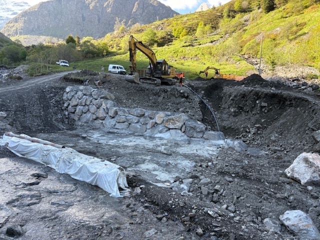 Pelle mécanique sur un chantier de terrassement en montagne avec enrochement et aménagement de cours d'eau.