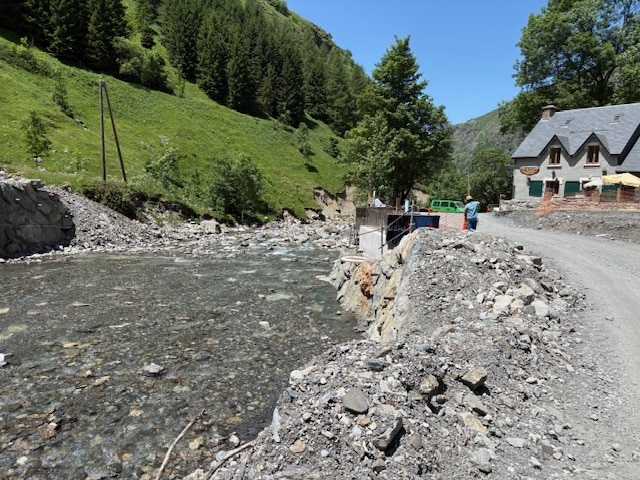 Torrent de montagne rocailleux, berges en travaux et auberge en pierre sous un ciel bleu ensoleillé.