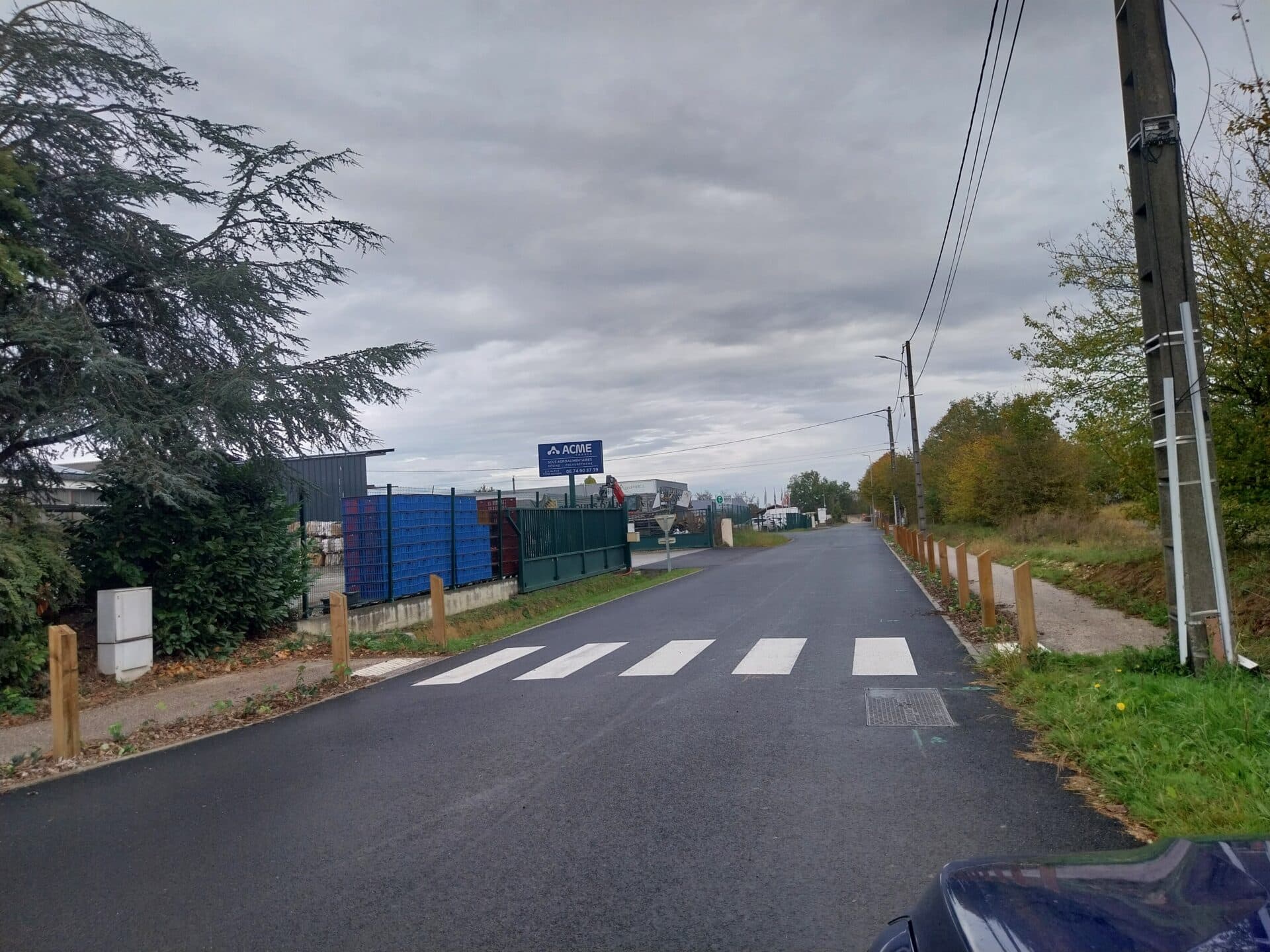 Passage piéton et panneau ACME à l'entrée d'une zone industrielle sous un ciel gris nuageux.