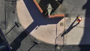 Ouvrier en gilet orange sur un chantier de voirie délimité, vue aérienne zénithale avec ombres.
